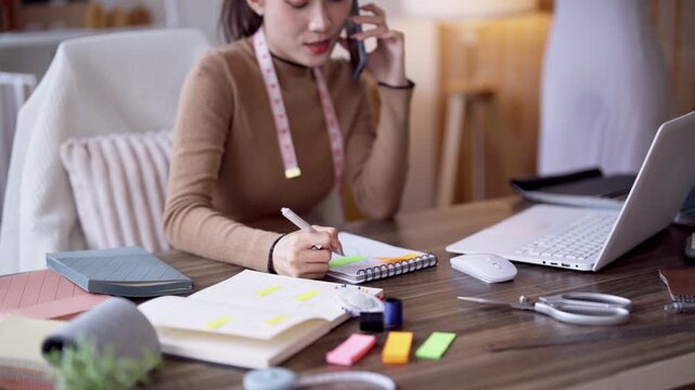Young Asian fashion designer talking on phone while taking notes at wooden desk in bright home studio