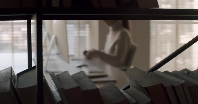 Out of focus businesswoman sitting at her desk, feeling stressed and anxious about work deadlines while books on a shelf are in the foreground, creating a sense of professional pressure