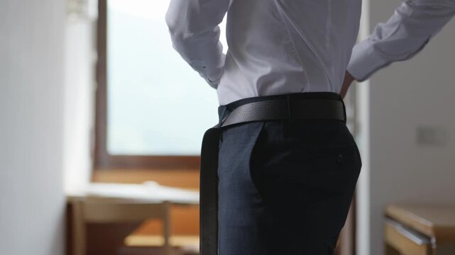 Groom getting dressed before wedding ceremony, fastening belt and preparing formal attire. Elegant pre-ceremony moment in a bright indoor setting.