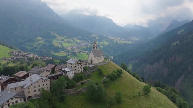 Aerial view of historic mountain church perched above Colle Santa Lucia in the Dolomites. Scenic alpine landscape with dramatic peaks, valley village and traditional architecture.