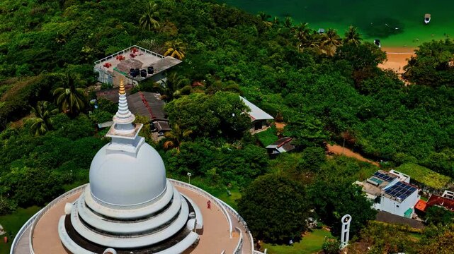 White Japanese Peace Pagoda crowns Rumassala hill as a drone flies past lush forest toward the small bay below.