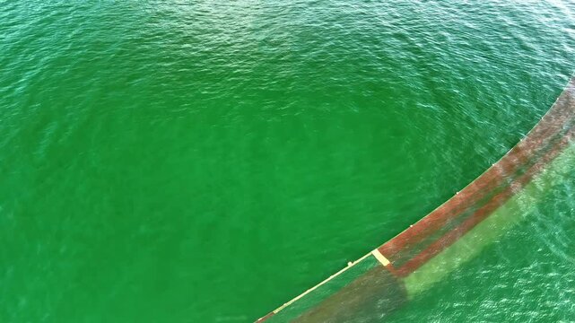 Large curved shore net arcs through bright green sea as a small fishing boat works near the top of frame, viewed from above.