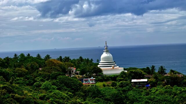Japanese Peace Pagoda rises over Rumassala hillside greenery with houses in the foreground and calm ocean under cloudy skies.