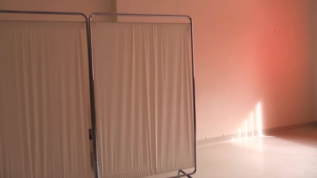 Quiet doctor's office with a medical folding screen and sunlight creating shadows on the floor and pink wall, creating a serene and empty atmosphere for healthcare and privacy concepts