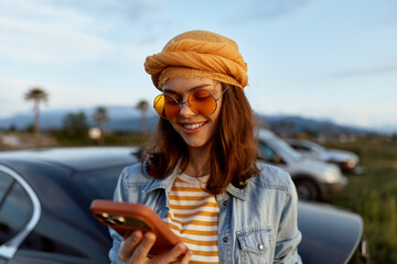 Obraz premium Smiling woman in denim jacket and striped shirt using a smartphone outdoors, with a car and distant hills in the background. Casual lifestyle scene highlighting travel, leisure, and personal moment.