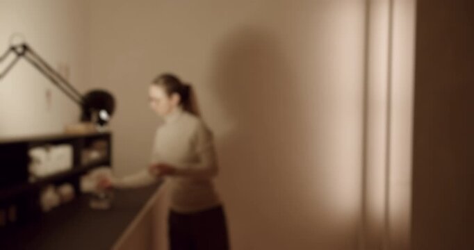 Out of focus woman with a ponytail finishing her work at a desk in a dimly lit office, turning around and walking away from her creative workplace in a cinematic, slow motion shot