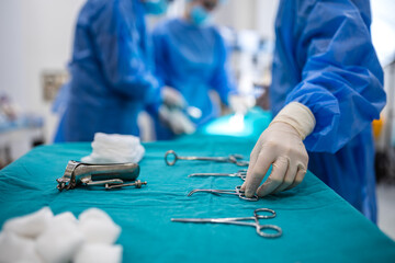 Medical professional in blue surgical scrubs and sterile gloves reaches for stainless steel surgical instruments on a tray in a sterile operating room environment during a planned surgical procedure. © Graphicroyalty