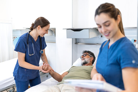 Caring female nurse in blue scrubs holds the hand of an elderly male patient resting in a hospital bed, while her colleague reviews his medical chart, providing compassionate care in a clinic
