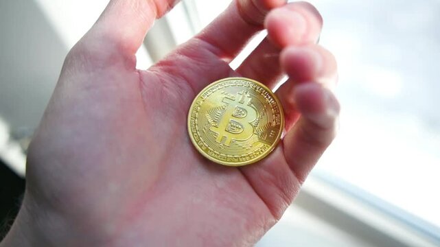 Human fingers holding shiny golden bitcoin coin against bright blurred background