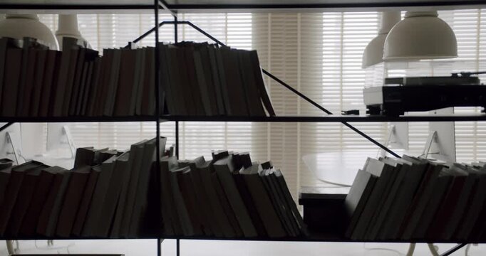 Dark modern bookshelf silhouette filled with numerous books and a vinyl record player standing against a window with closed blinds, revealing a sleek and stylish contemporary interior
