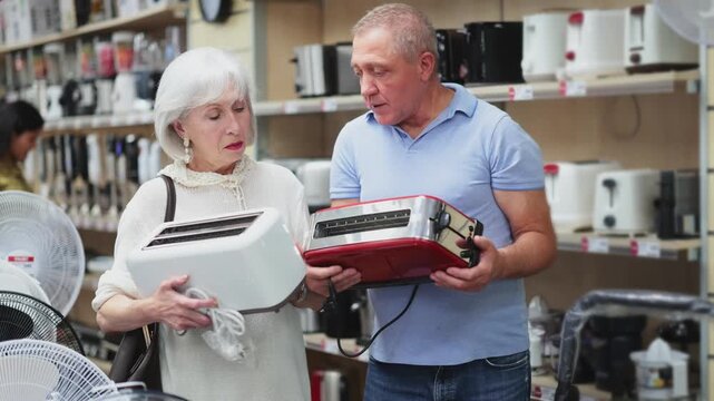 Elderly couple is standing in a store with appliances, they carefully choose a toaster to make bread for breakfast. Older husband and wife looking at toaster variations. High quality 4k footage