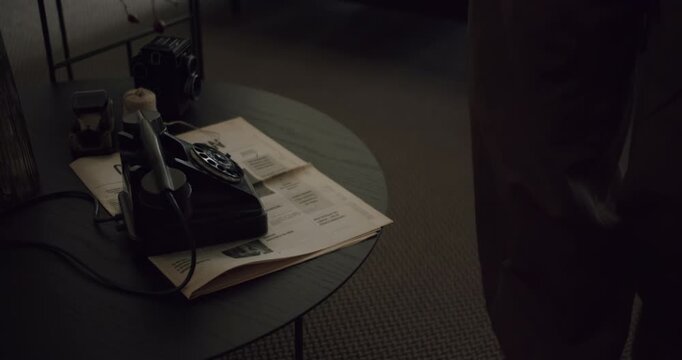 Low-angle slow-motion shot of a person's legs walking past a table with a vintage black rotary telephone, newspaper, and an old camera, creating a nostalgic and mysterious atmosphere
