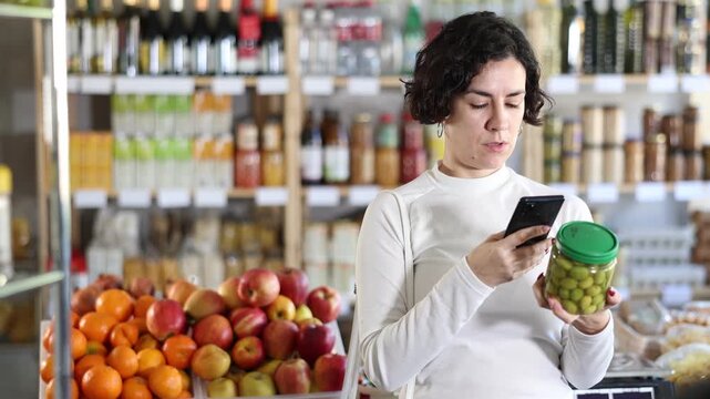Woman scans barcode QR code on pickled olives label package, follows link and studies information for consumer in supermarket application. Client seek olives manufacturer data in on-line marketplace. 