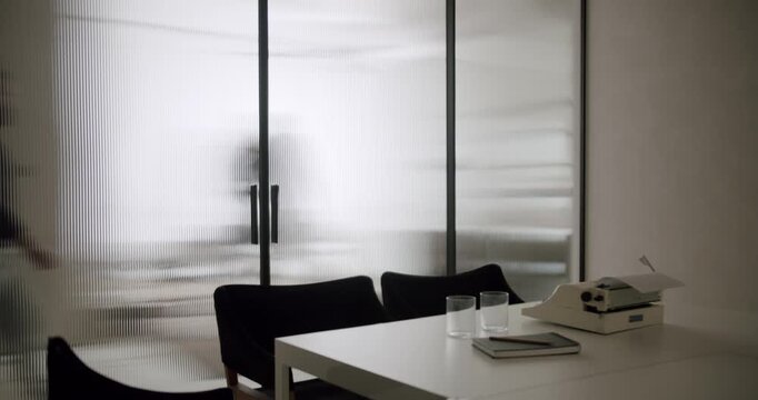 Silhouettes of people moving behind a translucent fluted glass wall in a contemporary office setting, with a minimalist desk, chairs, and a vintage typewriter in the foreground