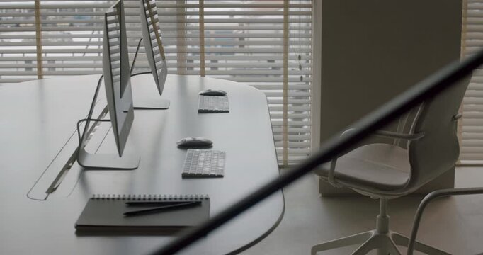 Minimalist and modern office interior showing empty workstations with computers, keyboards, and notebooks on a large desk by a window with blinds, creating a quiet and professional atmosphere