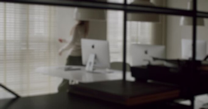 Defocused shot of a lonely woman walking thoughtfully through a modern, minimalist corporate office, pausing to look out the window before continuing on her way, evoking a sense of solitude