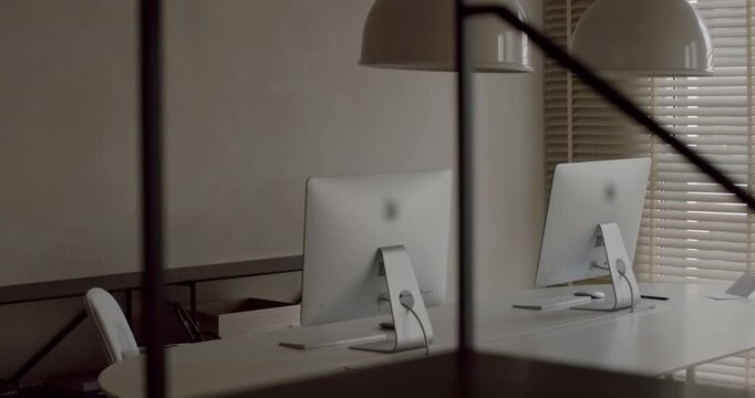 Empty minimalist office interior with white desks and computers, filmed through a blurred foreground of shelves. The contemporary and calm workspace is bathed in soft natural light