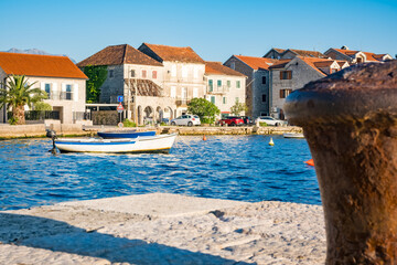 Mooring bollard in Tivat, Montenegro. A small white boat is anchored in the blue water. Colorful buildings line the shore under a clear sky. © Mila