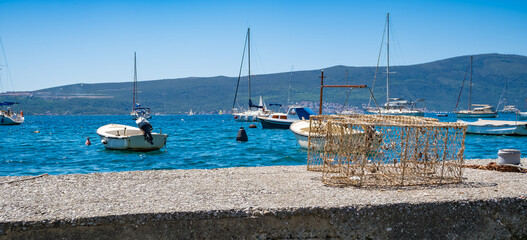 A crab trap sits on the shore near a yacht marina in Tivat, Montenegro. Several boats are anchored in the clear blue water under a bright sky. © Mila