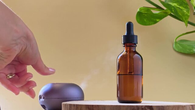 Slow motion hand pressing button on aroma diffuser blowing steam near amber bottle. Natural aromatherapy, essential oil wellness or spa routine concept.