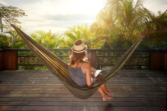 Young woman in straw hat relaxing in hammock on wooden terrace, reading book in tropical resort. Summer vacation concept.