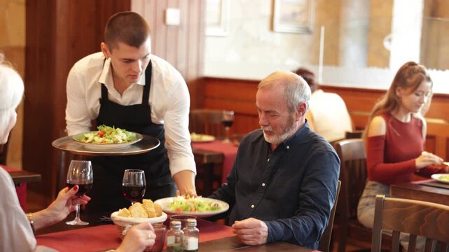 In restaurant hall, guy waiter brought order food to mature couple visitor, puts traditional Mediterrian salad of leafy greens, cheese on table. Waiter fetch and carry food for restaurant client