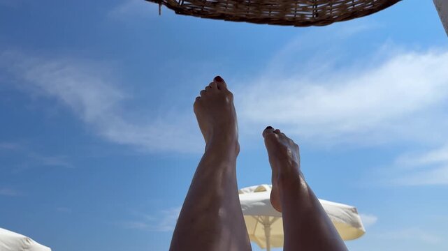 First-person POV perspective of slender female legs raised up towards the blue sky with clouds under a wicker beach umbrella
