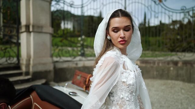 Beautiful woman in white vintage dress stands by classic car at elegant estate