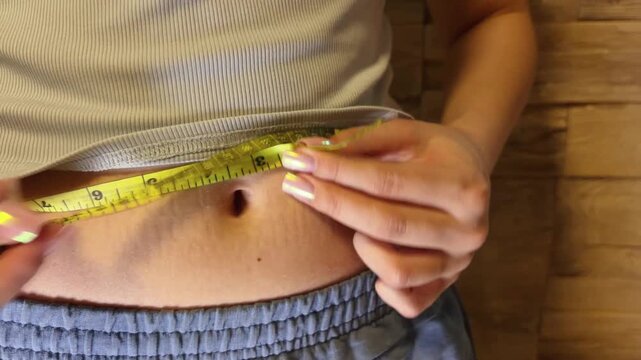 Slim woman measuring her waistline with a yellow tape, checking her body parameters for weight loss, diet, and fitness progress after postpartum recovery and workout