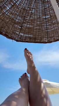 First-person POV perspective of slender female legs raised up towards the blue sky with clouds under a wicker beach umbrella