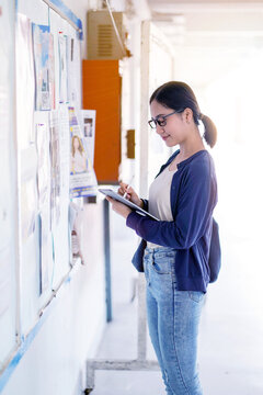 Happiness Asian girl, dressed in casual clothes, holds a tablet and takes notes on school announcements posted on the bulletin board in her school building.