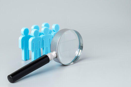 A magnifying glass in front of a group of blue human figures on a grey background. Recruitment, target audience analysis, human resources auditing concept.