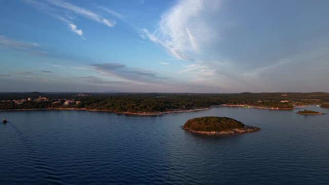 Small wooded island floats on calm Adriatic waters beside pine-covered Istrian coastline at golden sunset, with boat wake and wispy clouds near Vrsar, Istria, Croatia. Aerial drone wide shot.