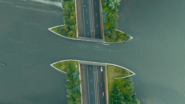 Aerial top-down shot of the Harderwijk highway aqueduct where cars pass beneath a water bridge. Lush green trees line the road, showcasing modern infrastructure blended with nature.