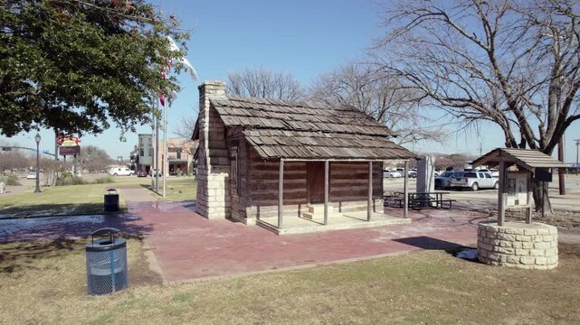 4K drone push-in footage of the historic Larkin Newton Log Cabin in downtown Midlothian, Texas, with American and Midlothian flags waving in the wind beside the landmark.