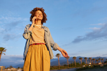 Obraz premium Young woman outdoors enjoying a sunny day, casual fashion with a denim jacket over a striped tee and a mustard skirt, a vibrant lifestyle portrait on an open coastal road.