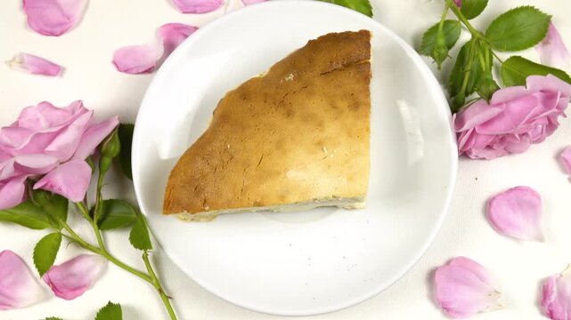 A white plate with a piece of homemade cottage cheese pie on it stands on a white table among roses and petals, top view