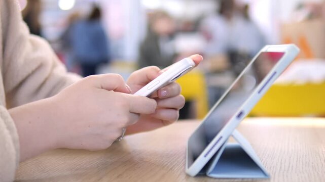 Hands hold smartphone, tablet nearby on wooden table. Person focused, calm, engaged in digital tasks. Suitable for tech, work-from-home, or multitasking themes. Background blurred, people out of focus