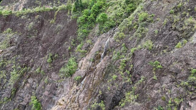Drone footage captured at Khanh Le Pass between Lam Dong and Khanh Hoa provinces, Vietnam. The drone flies directly toward a steep mountainside, documenting a narrow stream cascading down the rock fac