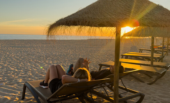 Beach at sunset with rows of sun loungers and straw umbrellas. The sky is painted in soft pastel shades of orange, pink and blue.
