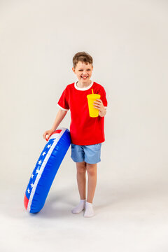 Little boy wearing a red t-shirt and blue shorts stands with an inflatable pool ring, holding a yellow drink, smiling cheerfully on white background. 4th of July pool party in USA