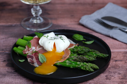 A dish features a poached egg on top of slices of ham and asparagus on a wooden table. A glass of drink is seen in the background. Healthy eating concepts. side view