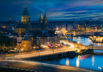 Obraz premium Stunning blue hour panoramic view of Amsterdam city centre with Basilica of Saint Nicholas and canals, illuminated by glowing city lights under a dramatic twilight sky, Netherlands