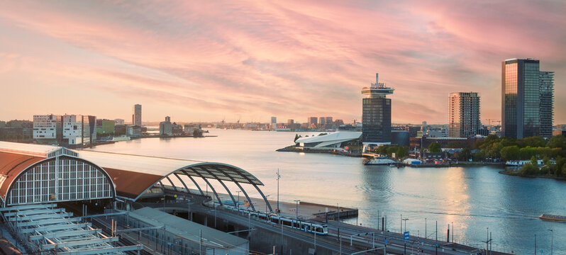 Panoramic view of Amsterdam Centraal Station, netherlands overlooking the IJ waterfront at golden hour featuring 'DAM Tower and EYE Filmmuseum illuminated by a colourful sunset.