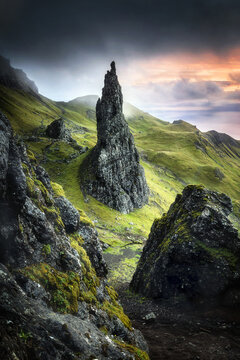 View of the Old Man of Storr rock pinnacle surrounded by green hills and dramatic clouds at sunset in Portree, Scotland, United Kingdom.