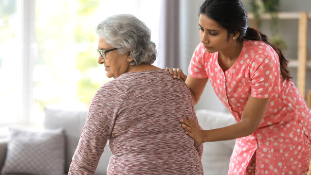 Young Indian nurse caregiver assisting and consulting an elderly mature woman with backache in a comfortable home environment, professional healthcare worker offering compassionate support.