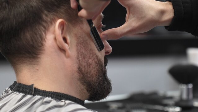 Professional barber performing precise beard shaving with classic straight razor, working in modern barbershop with skilled grooming technique