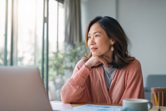Asian woman sitting at desk with laptop looking out window. Pensive girl brainstorming business ideas in home office. Professional planning future strategy. Remote work and lifestyle concept.