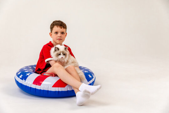 Little boy in red shirt and blue shorts stands sits in large inflatable pool ring with American flag print, holds ragdoll fluffy white cat in his arms, smiling at camera, white background, copy space