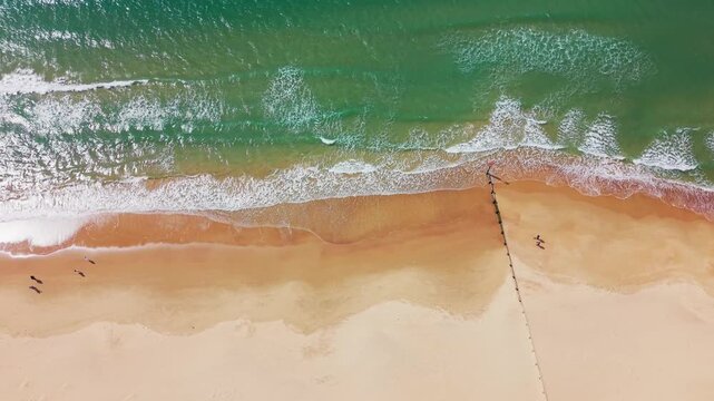 Top down aerial drone footage of waves washing onto a sandy beach with turquoise sea, white surf, shoreline patterns and a coastal groyne. Useful for themes of UK beaches, seaside travel, coastal land